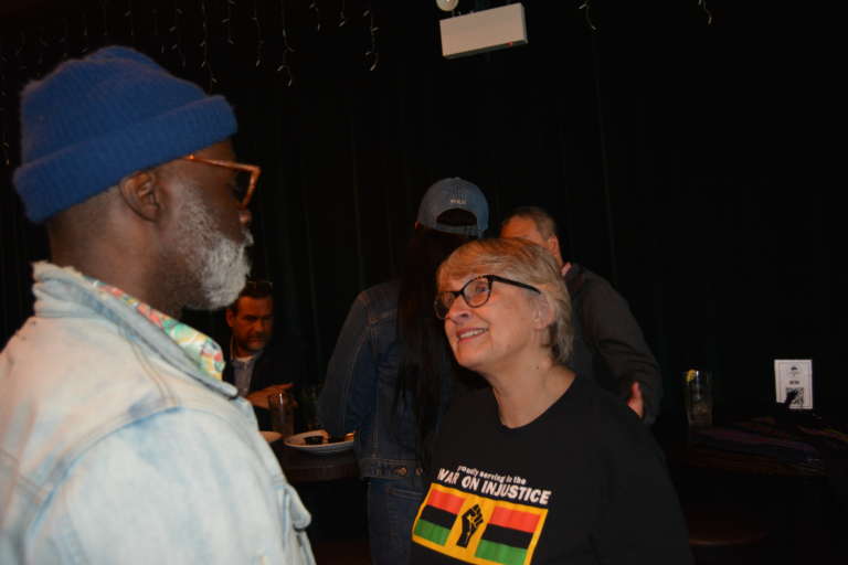 A black man wearing glasses and a denim jacket in conversation with a white woman in glasses and a black t-shirt