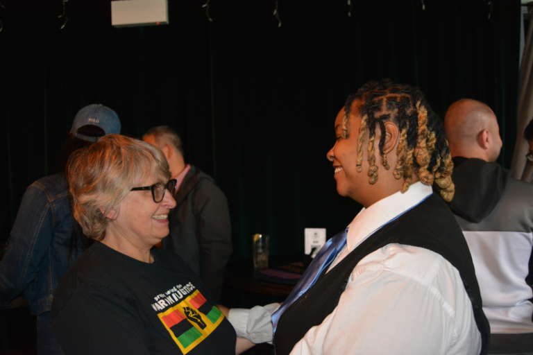 A white woman in a black T-shirt smiling and facing a Black woman in a collared shirt and vest who is reaching out to embrace her.