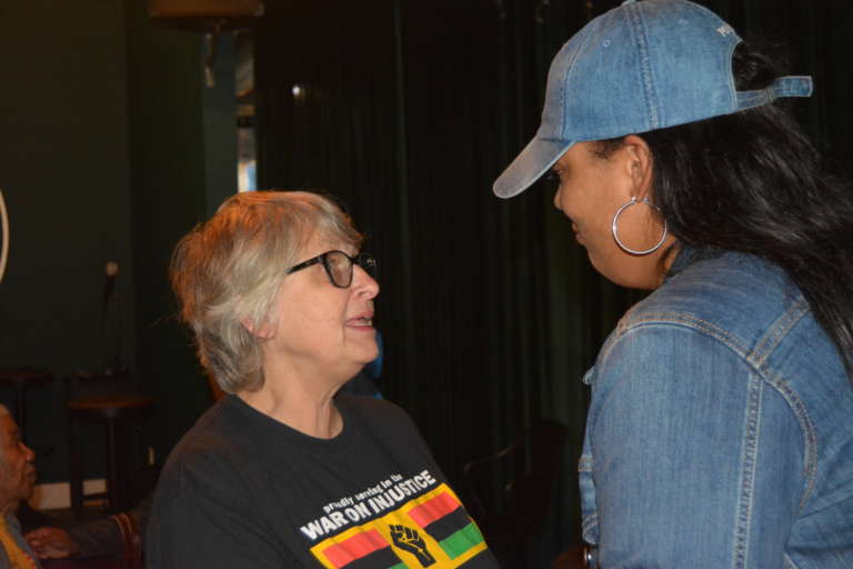 A white woman in a glass and a black t-shirt in conversation with a Black woman in a denim top and matching baseball cap