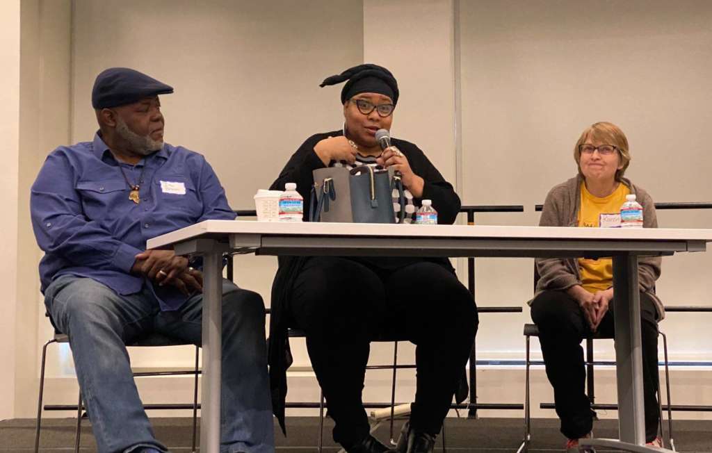 A panel table on a temporary stage in front of windows with pulled white shades. Three people are at the table. On the left is a Black man wearing a flat cap and turned to the middle, listening. In the middle is a Black woman in a black headscarf and glasses, holding a microphone and speaking. On the right is a white woman in a yellow t-shirt and glasses, reacting to what she's hearing.