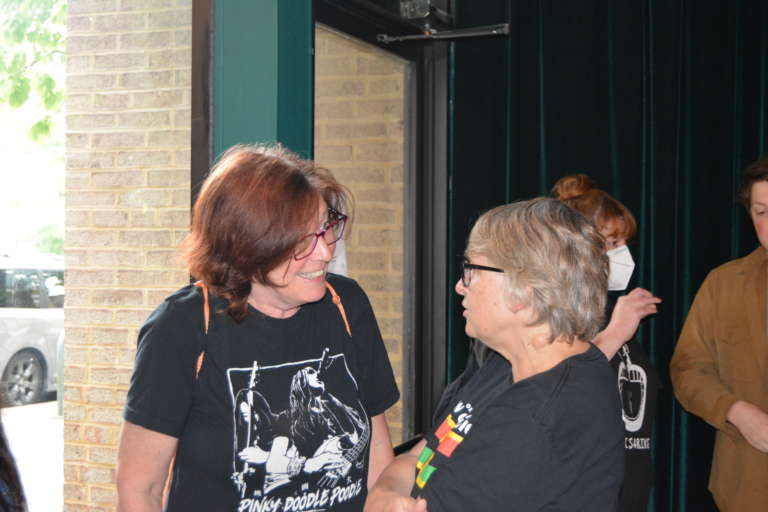 Two white women, both with glasses and wearing black T-shirts, in conversation with each other. Neither is facing the camera. A brick wall is visible outside a glass door and window.