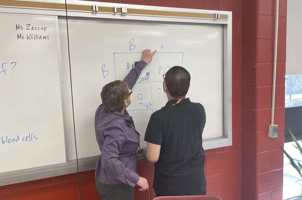 A classroom with red cinderblock walls. In front of a sliding white board, backs to the camera, are a white woman with a short haircut and purple blouse, showing work to a student with short dark hair and a black t-shirt.