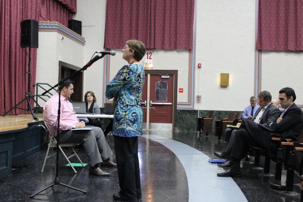 A school auditorium with burgundy curtains. In the foreground center of the photo, a white woman in a blue patterned tunic blouse is speaking at a microphone. Behind her, to the right of the photo, three men in suits sit in the auditorium chairs. To the left of her, in front of the stage, a man in a pink button down shirt shits in a folding chair facing the audience. Behind him, a woman in business casual attire sits behind a laptop at a folding table.