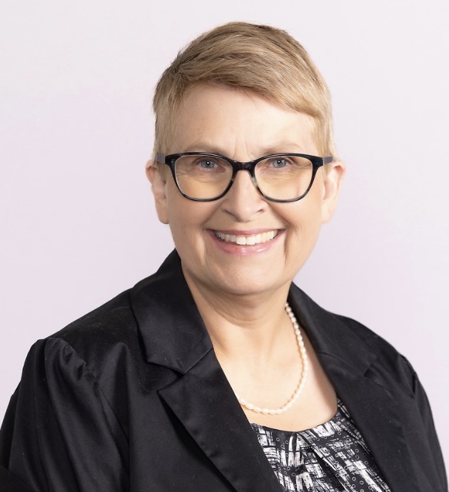 A white woman with short blond hair and glasses smiling while looking at the camera. She's wearing a dark blazer and a pearl necklace.